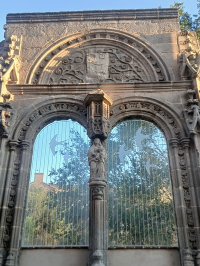 This image depicts the exterior of the ruins of the church of San Vicente in Ávila, Spain. The central focus is a large, arched stone structure that appears to be a window frame. The stonework is intricately carved with floral motifs and other decorative elements. Within the archway, there are two glass panels, each adorned with white silhouettes of birds in flight. These glass panels are set against a backdrop of trees and a building with a terracotta roof, visible through the glass. The overall impression is one of weathered historical architecture combined with modern artistic additions. The sunlight suggests it is either early morning or late afternoon, casting shadows and highlighting the textures of the stone. There are no people visible in the scene.
