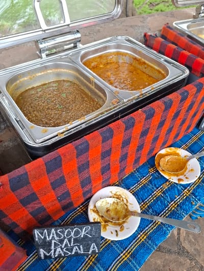 A food warmer, containing two compartments, is on a table covered in a checkered cloth. One compartment contains a light brown curry with a creamy texture. The other compartment contains a dark brown lentil stew with a more substantial texture. A single spoon, lying on a white plate, is to the right of the food warmer. There are two other spoons in the image, one of which is in a white bowl containing a light brown curry. The other spoon is on the ground. A black slate sign with the words "Masoor Daal Masala" is placed next to the plate. This scene takes place at a restaurant or food vendor in Karagita, Kenya, located at coordinates -0.773 latitude and 36.42 longitude.  There are no people present.