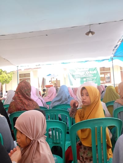 The image shows a group of people, mostly women and girls, seated in rows of green plastic chairs under a white canopy in the city of Kecamatan Kutawaringin, Indonesia. The chairs are arranged in rows, with the people facing a stage or podium that is out of frame. Some of the women are wearing colorful headscarves, while others are wearing plain scarves in muted colors. One girl in a yellow headscarf is seated in the middle of the image and is looking at the camera. The image was taken in a public setting, perhaps during a religious gathering or ceremony.  The coordinates provided indicate the image was taken in the city of Kecamatan Kutawaringin, Indonesia, but the exact location within the city is unknown.