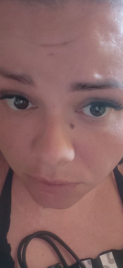 The media file is a close-up, vertically oriented photograph of a person's face and upper chest. The subject appears to be a young woman, with fair skin, dark eyebrows, and large, dark eyes. She is wearing what looks like dark clothing around her neck and shoulder. One of her eyes is clearly visible, looking directly at the camera. Her lips are slightly parted, and her skin has a smooth texture, with a mole visible on her cheekbone.
The lighting suggests an indoor setting, possibly with artificial light, as there are some shiny highlights on her skin. The overall mood is intimate due to the close-up framing. The background is largely out of focus, but the dark material around her neck, possibly a strap or collar, and the hint of patterned fabric on her shoulder are discernible. The image is candid and personal, focusing on the subject's features. There is no visible text in the image. The photograph is cropped tightly on the subject's face, with a portion of her neck and upper chest visible.