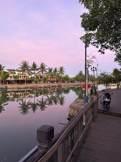 The image shows a peaceful riverside scene in Hội An Ward, Vietnam, during twilight. A calm river reflects the pastel pink and lavender hues of the sky and the row of traditional yellow buildings lining the opposite bank, interspersed with tall palm trees. Several small boats are moored along the river's edge. On the right side of the image, a wooden walkway runs alongside the river, bordered by a dark wooden railing. A black scooter is parked on the paved area next to the walkway, which is illuminated by decorative lampposts. The scene is serene, with no people visible in the frame.