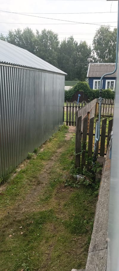 The image depicts a backyard scene in Krasnomayskiy, Russia, characterized by a narrow, unpaved path running between a corrugated metal structure on the left and a wooden fence on the right. The metal structure appears to be a shed or garage, with its ridged surface reflecting light. The wooden fence is weathered and consists of vertical planks, some of which are leaning or broken, with vegetation growing around its base. Beyond the fence, a blue house with white-framed windows is visible, partially obscured by dense green foliage forming an archway over another fence. The overall environment suggests a rural or semi-rural setting, with overgrown grass and trees in the background under an overcast sky. There are no people visible in the image.