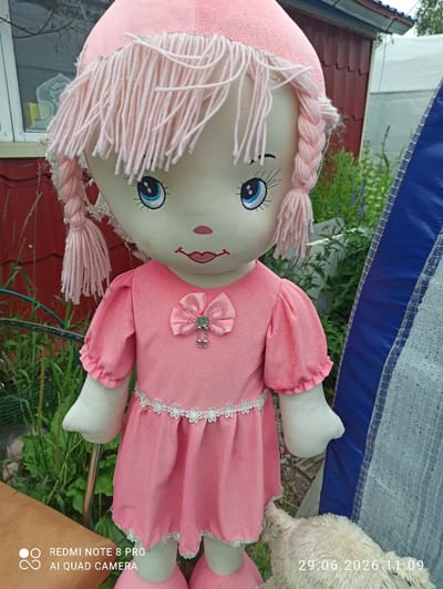 A life-sized stuffed doll wearing a pink dress and pink knitted hat with yarn hair is positioned outdoors. The doll has embroidered facial features, including blue eyes and a red mouth. A pink bow with dangling embellishments adorns the chest of the dress, which is trimmed with white lace. The doll is standing in front of a red building with a window and a white-framed greenhouse. Green foliage and a blue and white striped object are also visible in the background. The timestamp "29.06.2026 11:09" is present in the lower right corner, suggesting the image was taken on this date and time. The watermark "REDMI NOTE 8 PRO AI QUAD CAMERA" indicates the device used to capture the photograph. The location is identified as Sovetskiy, Russia.