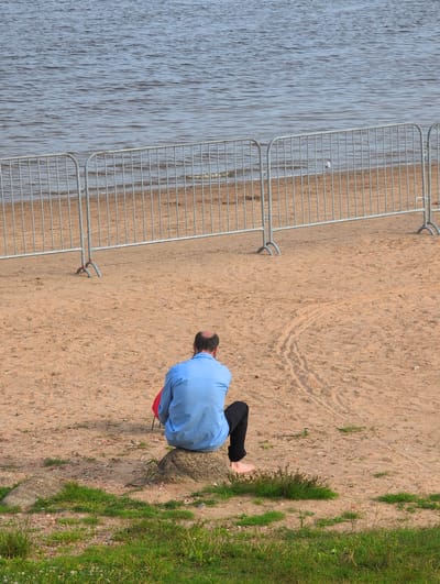 A man wearing a blue shirt and black pants sits on a rock on a sandy beach. He is facing away from the camera. In the background, a metal fence separates the beach from the water. The water appears calm with ripples. There are tire tracks in the sand behind the man. The scene is likely located near the Volkhov River in Veliky Novgorod, Russia, given the characteristic sandy banks and the presence of a barrier fence often used for events or public access control along the riverfront.