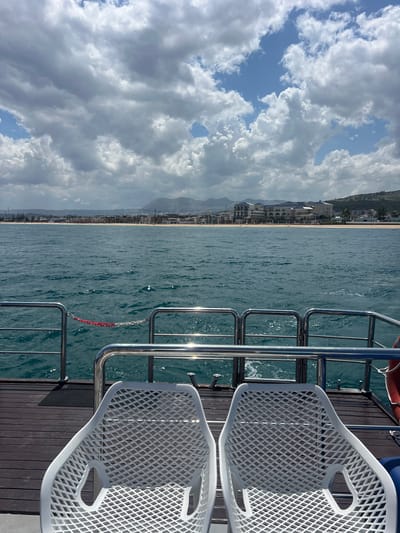 The photograph presents an elevated perspective from the deck of a vessel, presumably a boat or yacht, near M'Diq, Morocco. The image frame is divided vertically, with the right side focusing on elements of the boat. Two white, plastic, woven chairs are visible, positioned on a dark wooden deck. Behind the chairs, stainless steel railings reflect sunlight, indicating a water body.
To the left, the photograph reveals a coastal scene. M'Diq's shoreline is lined with buildings. A beach extends along the coast, with the city receding into distant mountains partially obscured by a cloudy sky with patches of blue. The water body appears relatively calm.