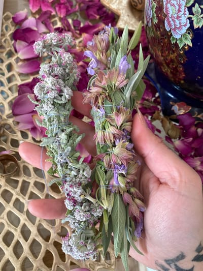 A close-up shot presents a person's hand holding two tied bundles of herbs, likely sage. The upper bundle features green leaves and pale purple flowers. The lower bundle displays smaller, gray-green leaves with tiny white and pinkish-purple flowers. Both bundles are bound with light blue string. The hand holding the bundles has a tattoo visible on the wrist. The background includes scattered rose petals, a decorative blue and gold ceramic object, and a gold, perforated surface. The location, Kunje, Montenegro, is not evident from the visual elements alone.