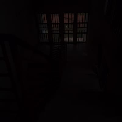 The image shows a dimly lit interior space, likely a building. The most prominent feature is a large window with multiple panes, each divided by horizontal and vertical bars. Through the window, some muted outdoor scenery is visible, suggesting it's daytime outside, although the interior is dark. The bars on the window create a grid-like pattern.
There is a sense of depth, with what appears to be a staircase or ramp leading downwards from the right side of the frame. Shadows are cast extensively across the floor and walls, obscuring many details. The overall impression is one of an enclosed, perhaps utilitarian or institutional space.
Given the location in Ogbomosho, Nigeria, this could be a part of a school, a church, a public building, or even a residential property with security features. The barred window might indicate a need for security or a stylistic architectural choice common in certain regions. The darkness suggests the photograph was taken during a time of low light, possibly early morning, late evening, or in an area with limited artificial lighting. There are no people or clear actions depicted, making it a still, observational shot of the environment.