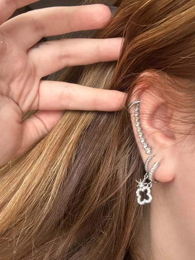 This close-up shot captures a partial view of a person, focusing on their right ear, strands of hair, and a hand. The person has long, wavy hair, primarily dark brown with prominent lighter blonde or caramel highlights, creating a streaked effect.
Adorning the ear are multiple silver-toned earrings. A distinctive ear cuff or climber, studded with small, clear rhinestones, follows the curve of the outer ear cartilage. Below it, a small, simple silver hoop earring is visible in the lobe. Further back, a dangle earring hangs, featuring a four-leaf clover or flower-shaped charm also encrusted with clear stones, and a smaller star-like charm attached.
A light-skinned hand, likely the person's left hand, is positioned in the foreground, with its fingers gently pulling back or holding strands of hair, revealing the ear and its adornments. The scene appears to be an intentional pose to showcase the hair and earrings.
The lighting suggests an indoor setting or a brightly lit outdoor area, but the specific time of day is indeterminate. There are no explicit background details to indicate a particular location within Troyan, Bulgaria, beyond the provided context. No visible text is present in the image, and emotions are not discernible from the visible portions of the subject.
