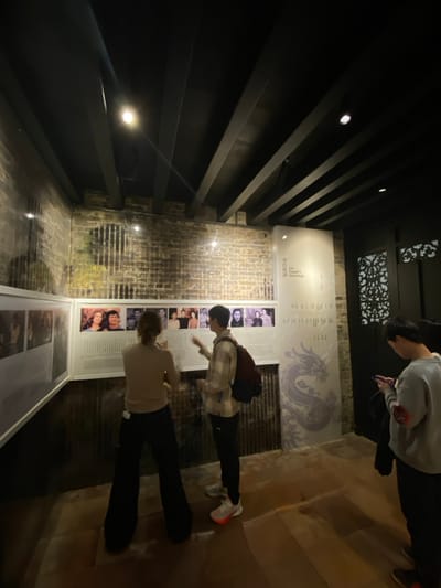 This media file depicts an indoor scene within what appears to be a museum or exhibition space, likely located in Guang Zhou Shi, China. Three individuals are present, observing various display panels.

The setting features a ceiling composed of dark, exposed wooden beams with integrated spotlights, and decorative patterned panels are visible towards the upper back of the room. The floor is made of polished wood, reflecting some of the overhead lighting.

Along the right side of the image, several display panels are arranged. A large white panel at the back features a subtle light purple dragon motif, Chinese characters, and partial English text that appears to say "On Family V... Genealogy." Below this, a series of smaller white panels present rows of black and white photographs of individuals, each accompanied by descriptive text.

In the mid-ground, a person wearing a plaid shirt and dark pants is leaning forward, engrossed in an exhibit. In the foreground, another individual wearing a light-colored top and dark pants is also focused on the displays. Further back, a third person in a light grey hoodie stands, seemingly interacting with a mobile phone.

The left wall of the room has a textured surface, possibly brick or distressed plaster, which is illuminated by spotlights. The overall lighting is artificial and somewhat subdued, creating a focused atmosphere for viewing the exhibits. The people appear calm and engaged in their activity.