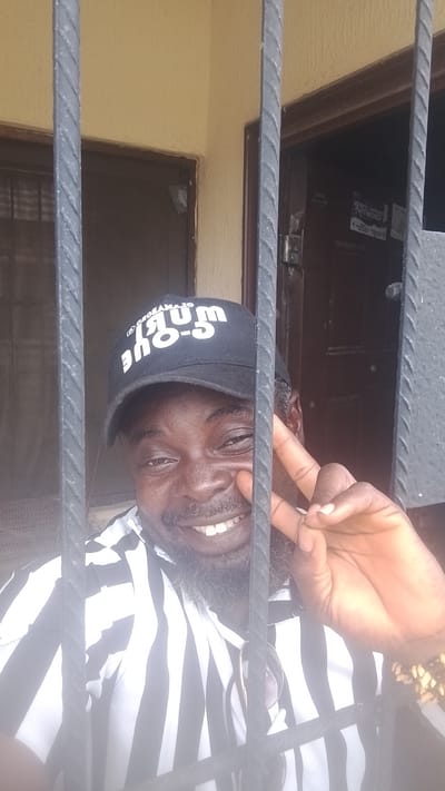 In Asubi, Nigeria, a Black man with a beard and mustache, wearing a black baseball cap with white lettering that reads "C-QUEUM," and a black and white striped shirt, smiles and makes a peace sign with his right hand. He is looking at the camera through black metal bars, which create a visual barrier in the foreground. Behind him, a doorway with two wooden doors is visible, and a portion of a light yellow wall with some texture is to the left. There are also some papers taped to the right door. The man has sunglasses hanging from the collar of his shirt. He is wearing a beaded bracelet on his right wrist.