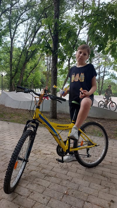 In a park setting in Lermontov, Russia, a young boy with blond hair stands next to a bright yellow bicycle. He is wearing a black t-shirt with a cartoon cat design and black shorts. He is holding a small, light-colored snack in his right hand and a plastic wrapper in his left. The bicycle is a "DAKOTA" model, featuring blue accents and "FORWARD" branding. The boy's left foot is resting on a pedal, and his right foot is on the ground.
In the background, another person is visible riding a bicycle on a paved surface that appears to be a skate park or a similar recreational area. The ground is paved with light-colored bricks or tiles, and dry grass and leaves are scattered on the surface. Tall trees with lush green foliage surround the area, suggesting a warm, possibly summer, day. The overall mood seems relaxed and casual, implying a leisure activity. The lighting suggests it is daytime.