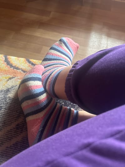 The image shows a close-up view of a person's feet, wearing striped socks and purple pajama bottoms. The socks have a pattern of pink, blue, white, and black stripes. The person's feet are resting on a textured surface, possibly a couch or blanket, which has a geometric pattern in shades of yellow, orange, and gray. In the background, a wooden floor is visible, with warm lighting suggesting an indoor setting. The scene appears to be casual and relaxed, perhaps during the day given the lighting. There is no visible text in the image.