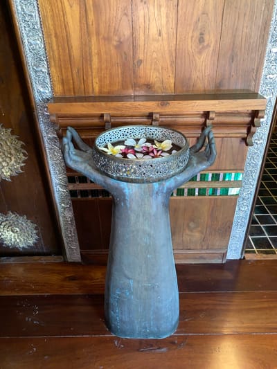 The image shows an ornate bronze statue of two hands cupped together, supporting a decorative metal bowl filled with water and plumeria flowers. The statue stands on a wooden floor in front of a large wooden door adorned with intricate silver detailing on the left and right sides. The door has vertical wood paneling. Above the statue is a wooden shelf. To the right of the statue, a wall features a decorative strip of colorful mosaic tiles. The overall scene suggests an interior space with a rich, cultural aesthetic, possibly a hotel lobby or a traditional Thai home in Baan Lamai, Thailand.