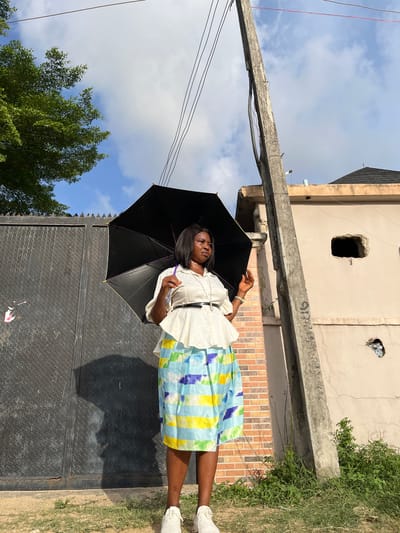 An adult female stands outdoors in Iranla, Nigeria, holding an open black umbrella overhead. She is positioned facing slightly right of the viewer, looking towards her left. She wears a white blouse with a black belt, a knee-length skirt patterned with light blue, yellow, and darker blue stripes, and white athletic shoes. Her shadow, including that of the umbrella, is cast clearly onto a large, dark gray diamond-plate metal gate to her immediate left. The gate features spiked anti-climb elements at its top. To her right stands a weathered concrete utility pole, exhibiting cracks and supporting multiple dark electrical wires overhead. A light-colored building with a dark, possibly tiled roof is visible in the background, to the right of the pole. This building features a large, irregular rectangular opening at mid-height and a smaller, similar opening lower down, containing reflective debris. A section of a brick wall is visible between the gate and the utility pole. The ground consists of sparse dry grass, dirt, and green weeds. A green deciduous tree is partially visible on the far left, under a partially cloudy blue sky.