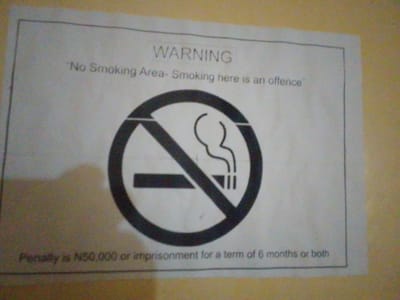 A printed sign is displayed on a light-colored wall, possibly indoors or under a covered outdoor area, in Nyanya, Nigeria. The sign is framed by a thin black border. At the top, the word "WARNING" is printed in bold, uppercase letters. Below this, a sentence reads, "No Smoking Area- Smoking here is an offence". The central and largest element of the sign is a circular graphic depicting a crossed-out cigarette. Inside the circle, a stylized cigarette with smoke rising is shown, with a thick black diagonal line passing through it. Below the circular graphic, a sentence states, "Penalty is N50,000 or imprisonment for a term of 6 months or both". The lighting in the image is uneven, casting shadows and creating a slightly grainy appearance. The wall behind the sign is a warm, yellowish-brown color.