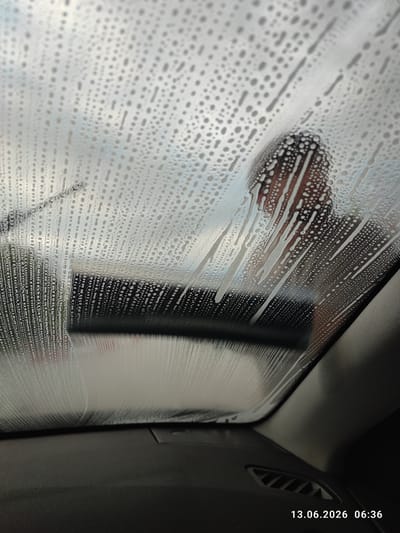 The image shows a close-up view of a car's windshield covered in water droplets and soap suds. A person's head and shoulders are visible in the upper right quadrant of the image, reflected in the soapy windshield. The person appears to be a male with dark hair. The windshield wiper is visible on the left side of the image, suggesting it is in use or has recently been used. The interior of the car, including the dashboard and an air vent, is visible in the lower part of the image. The timestamp "13.06.2026 06:36" is visible in the bottom right corner, indicating the date and time the photo was taken. The setting appears to be a car wash or a location where the car is being cleaned. The provided metadata indicates the image was taken in Belozem, Bulgaria.