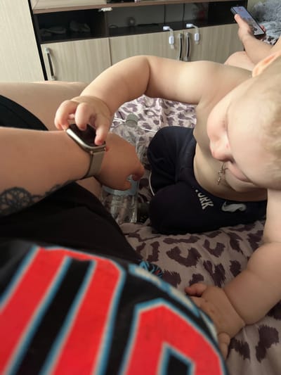 The image shows a close-up, overhead view of a baby and an adult's arm. The baby is wearing black sweatpants with "YORK" printed on them in white letters. The baby is looking down at something held in their hand, which is out of focus. The adult's arm, also in focus, is wearing a silver watch on the wrist and has a tattoo on the forearm. The adult is holding what appears to be a smartphone. Both subjects are lying on a bed with a leopard-print comforter. In the background, part of a dresser or cabinet with light-colored drawers and silver handles is visible, suggesting an indoor setting. Several plastic water bottles are also visible on the bed. The lighting suggests it is daytime. The context provided is Chelyabinsk, Russia.