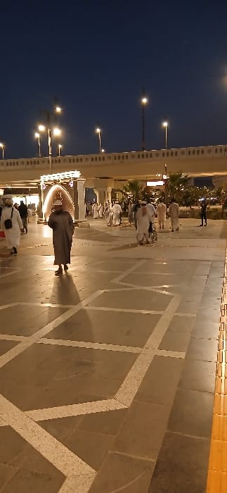The image depicts an urban pedestrian area at night in Medina, Saudi Arabia, specifically in the vicinity of Al-Masjid an-Nabawi. The scene is dominated by a wide, tiled walkway featuring large, light brown/grey squares interspersed with geometric patterns in lighter beige.

In the foreground and midground, numerous individuals are present, predominantly adult males dressed in traditional Saudi attire, including white or dark-colored thobes and various head coverings such as ghutras. One individual in a dark thobe and light head covering walks towards the viewer in the center. To the left, a person in a white thobe with a black shoulder bag walks away. Towards the right midground, several individuals are moving, with one appearing to push a small wheeled cart or wheelchair.

Behind the pedestrians, a prominent illuminated arched structure, possibly a gate or decorative entrance, glows brightly. Further in the midground, beyond the archway and to the right, landscaped areas with green foliage and additional lighting are visible.

The background features a large, light-colored concrete overpass or bridge spanning the width of the image. The bridge's railing exhibits decorative cutouts or patterns. Numerous tall streetlights with bright lamps illuminate the bridge and the area beneath it, casting a glow against the dark night sky. The overall environment is well-lit and appears to be a busy public space.