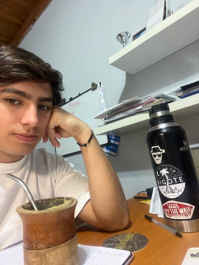 A young man is posing for a selfie. He is sitting at a desk with a gourd (mate cup) with a silver straw (bombilla) in the foreground. A black Stanley thermos with stickers including "Bigote" and "Vans Off the Wall" is on the desk. Also visible on the desk are a pen, papers, and a coaster. A white shelf is above the desk. Based on the poster with the name "Rocco VIllaveiran" visible in the background, this image was taken in Gerli, Argentina.