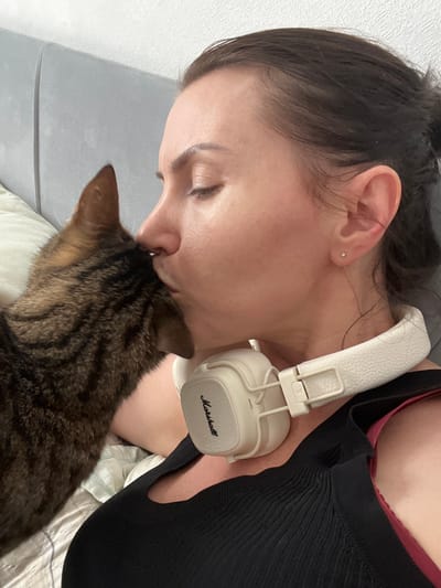 A close-up, waist-up photograph shows a person with dark hair pulled back, leaning against a grey headboard in a bed. A tabby cat is nuzzling the person's face, with its head against the person's cheek and nose. The person is wearing white headphones around their neck, with the brand name "Marshall" visible. They are also wearing a black top. The background is a textured grey wall and a white pillow. The scene appears to be indoors, likely in a bedroom. The specific location in the city of Tolyatti, Russia, cannot be determined from the image itself.