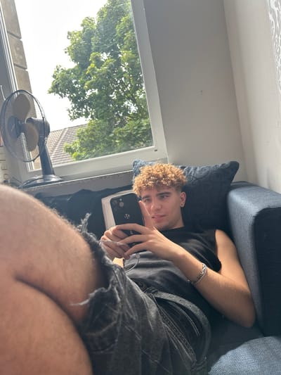 The media file shows a young man with curly blonde hair lying on a blue sofa, looking at the camera. He is wearing a black t-shirt and dark denim shorts. He has a silver bracelet on his left wrist. He is holding a black smartphone in his right hand, which is positioned in front of his face. The background shows a window with trees and a clear sky, suggesting it is daytime and the weather is pleasant. A black electric fan is visible on a windowsill below the window. The overall setting appears to be a residential indoor space. There is no visible text in the media file. The image is captured from a low angle, with the camera tilted. The man's expression is relaxed and he seems to be posing for the picture. The lighting suggests it is a sunny day.