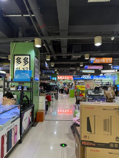 This is an indoor shot, likely inside a computer and electronics market or mall. The perspective is looking down a narrow aisle lined with various vendor stalls. Banners and signage hang overhead, with Chinese characters and English text visible. One banner advertises "Meili Mobile Phone Shop 2C30" (美丽手机铺 2C30) and another mentions "Zhongguancun Laptop Repair". Other store signs include "Duodu" (多多) and "NanJing Guomei Digital" (南京果美数码).
Several boxes, including one for a "Great Wall M69 Pro" computer case, are stacked in the aisle. The environment is cluttered with electronics, computer parts, and packing materials. There are some individuals visible in the background and a person wearing a mask and looking at a phone on the right side of the image.
The location is confirmed by one of the vendor signs, which includes the phrase "Xuanwu District Yinuo Electronics Operating Department" (玄武区一诺电子经营部), indicating it is within Xuanwu District in China. The lighting is a mix of fluorescent overhead lights and spots.