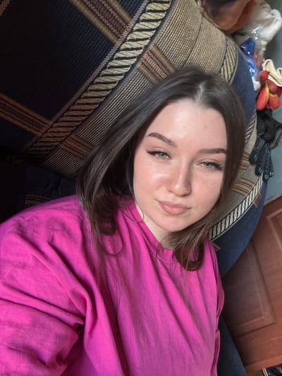 A young woman with brown hair and green eyes is captured in a selfie, lying on a couch. She is wearing a bright pink t-shirt and appears to be indoors, with a wooden door visible in the background. The couch has a patterned fabric in shades of blue, brown, and beige. Some objects are visible behind her head, including what appears to be a white plastic bag and some red and yellow items that could be gloves or small toys. The lighting suggests it might be daytime, but no specific time or weather conditions can be determined. The woman's expression is relaxed and she has a gentle smile. The overall scene evokes a casual and comfortable atmosphere. No visible text is present in the image.