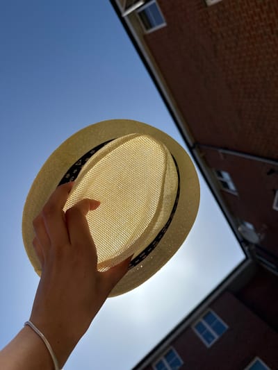 A low-angle shot shows a straw fedora hat held by a hand against a bright blue sky, partially obscuring the sun. The hat has a black patterned band around the brim. The hand is light-skinned and adorned with a silver bracelet. In the background, blurred brick buildings with windows flank the sky. The image appears to be taken in an urban environment, potentially Great Yarmouth, United Kingdom, due to the architectural style of the buildings.