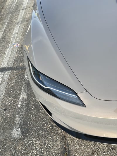 This is a close-up, angled, outdoor, eye-level shot of the front passenger side of a light brown Tesla car. The car is parked on an asphalt surface with faded white parking lines. To the left of the car, on the ground, is a small pile of colorful debris. The car's headlight is clearly visible, as is the edge of the hood and front bumper. The lighting suggests it is daytime, with sunlight creating reflections on the car's surface. The background is out of focus, but appears to be a street or parking lot setting. There are no people or other vehicles visible in the frame. The scene is located in Westminster, United States of America.