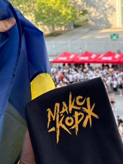 The image shows a close-up view of a hand holding a black fabric with yellow lettering, along with the corner of a blue and yellow flag, likely the Ukrainian flag. The lettering on the black fabric appears to be a stylized logo or text. In the blurred background, a crowd of people can be seen, some holding red banners with white lettering, possibly at an outdoor event. There are also red and white striped structures in the background, which might be tents or flags. The scene suggests a public gathering or event. The lighting indicates it is daytime, likely late afternoon or early evening given the warm tones. There are trees and greenery visible in the upper left portion of the background, suggesting an outdoor setting, possibly in a park or stadium area. A green sign with a white symbol is visible on a light-colored wall in the distance.