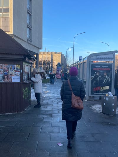 This vertical daytime photograph depicts a sunny street scene in an urban environment, likely Kraków, Poland. The sky is bright and clear blue, with faint contrails.
In the mid-ground, a person is seen from behind, wearing a dark puffy winter coat, a purple knit beanie, and a brown leather shoulder bag. They stand facing a modern bus stop shelter. The shelter features a transparent panel displaying numerous bus route numbers, including "109, 124, 134, 144, 164, 169, 173, 179" and "194, 300, 301, 304, 307, 310, 469, 424, 494, 502, 503," alongside the names "FIGURSKI KĘDZIERSKI." An advertisement on the shelter prominently features the words "DESPERANOS" and "ANTYRADIO" with two dark, stylized figures. A grey trash bin, surrounded by patches of snow or ice, stands next to the bus stop.
In the foreground, another individual wearing a light-colored coat and a hat leans over a brown kiosk, which is adorned with various newspapers and magazines. A white sign on the kiosk clearly states "W SPRZEDAŻY OBWARZANKI" (Obwarzanki for sale), hinting at the local delicacy. The kiosk also has some graffiti on its side.
The ground is paved with large, rectangular grey tiles, and a small pink piece of paper or litter is visible on the sidewalk near the person in the dark coat. In the background, several buildings with windows and architectural details are visible, along with two distinct streetlights curving into the sky. Other blurry figures suggest the presence of more people further down the street. The scene captures a moment of daily activity at a city bus stop and newsstand during a cold but sunny season.