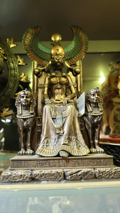 This media file showcases a close-up, front-facing view of an ornamental statue depicting an ancient Egyptian deity. The central figure, seated on a throne, has the head of a lioness and wears a headdress with a solar disk and uraeus. She is adorned with elaborate jewelry, including a wide collar, armbands, and bracelets. Her chest is bare, and she is clad in a draped, flowing robe. In her lap, she holds an ankh, a symbol of life.
Flanking the central figure are two sphinx statues, each with the head of a human and the body of a lion, facing forward. The sphinx on the left has a distinct nemes headdress, and both are depicted with intricate details on their chests. The base of the statue is decorated with Egyptian hieroglyphs. The overall material appears to be a metallic alloy, with a bronze or gold-like finish.
The statue is set against a blurred background of what appears to be a display or shelf, with other objects and lights visible, suggesting it is an artifact in a museum or a shop. The lighting is bright, casting some reflections on the polished surfaces of the statue. There is a small piece of white paper with some text attached to the right side of the central figure, though the text is mostly illegible. The setting is indoors, possibly in Vyborg, Russia. The statue is presented as a static display, with no discernible activity taking place.