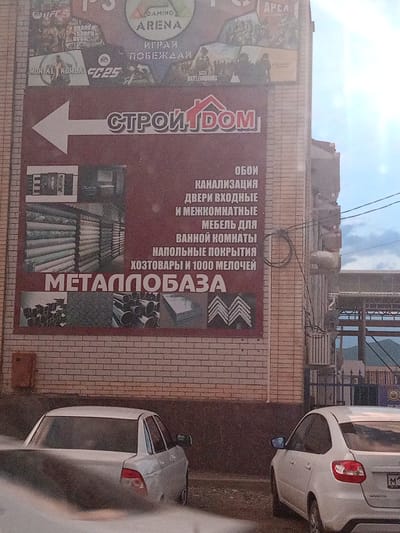 The image shows a brick building with a large advertisement on its side. The advertisement features the text "СТРОЙДОМ" (Stroydom) prominently, accompanied by an arrow pointing left. Below this, smaller text lists various building materials and home goods such as "ОБОИ" (Wallpaper), "КАНАЛИЗАЦИЯ" (Sewerage), "ДВЕРИ ВХОДНЫЕ" (Entrance Doors), "МЕБЕЛЬ ДЛЯ ВАННОЙ КОМНАТЫ" (Bathroom Furniture), "НАПОЛЬНЫЕ ПОКРЫТИЯ" (Floor Coverings), "ХОЗТОВАРЫ И 1000 МЕЛОЧЕЙ" (Household Goods and 1000 Small Items), and "МЕТАЛЛОБАЗА" (Metal Base). Images of wallpaper rolls, doors, metal pipes, and construction materials are also displayed. The building appears to be located in Zelenchukskaya, Russia, as indicated by the presence of a white car with a license plate starting with "M1". A second white car is partially visible in the foreground. The sky is overcast.