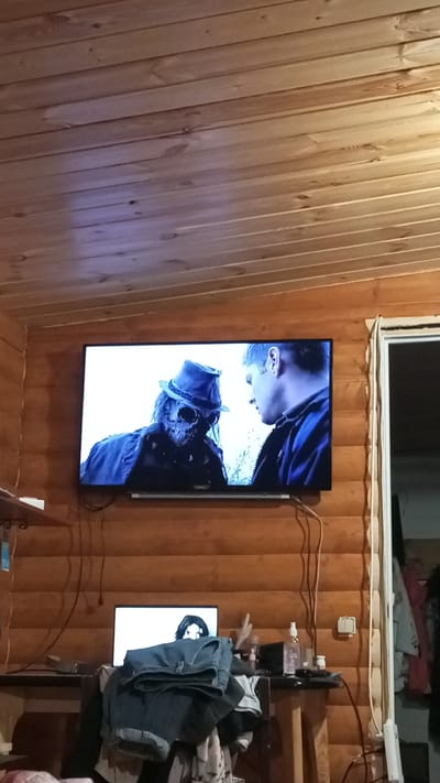 A dimly lit interior space with wooden walls and ceiling features a mounted television screen displaying a scene from a movie or show. In the scene, two characters are visible: one in a costume featuring a hat and mask with skeletal features, and another in a dark jacket. Below the TV, a desk cluttered with various objects, including a laptop, clothes, and bottles, is present. To the right of the TV, a partially visible doorway or window can be seen. The environment appears to be a living space. No people appear in the physical space of the image. The location is Юдино, Russia.