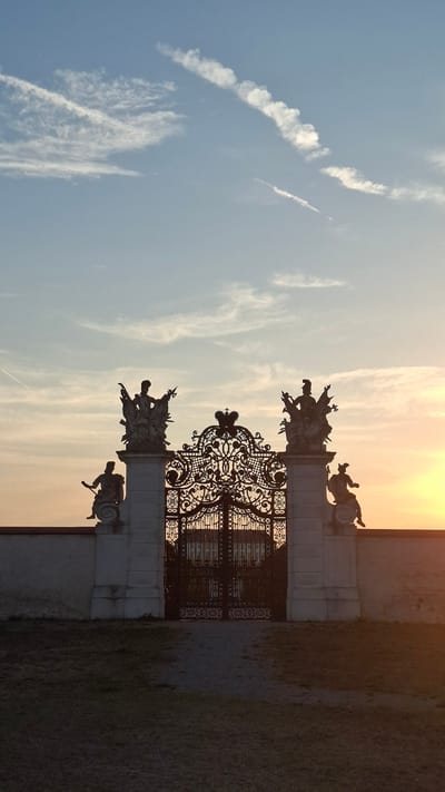 The image shows a silhouetted ornate gate at Schloss Hof, Austria, during sunset. The gate is flanked by white stone pillars topped with statues, and a decorative ironwork structure, possibly a crest, is visible above the gate. The sky is a gradient of blue and orange, with wispy clouds and contrails from an aircraft. A low wall runs horizontally across the frame, separating the foreground from the gate and sky. The ground in front of the gate appears to be a path with patches of dry grass. The overall scene is serene and captures the grandeur of the estate at dusk.