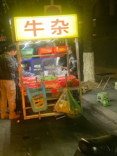 The image captures a vibrant street food scene at night in Guangzhou, China. A mobile food cart, brightly lit, is the central focus, set against a dark background.

The cart features multiple shelves holding red and green plastic baskets, likely filled with fresh ingredients. Steam rises from behind these baskets, indicating food preparation. Inside the cart, a vendor wearing a dark top is actively engaged in cooking. A large, illuminated yellow sign atop the cart displays the Chinese characters "牛杂" (Niúzázá), suggesting the stall specializes in beef offal or a variety of beef dishes.

Standing to the left of the cart is a man in a dark jacket and light brown cargo pants, looking towards the stall, possibly a customer or a fellow worker. Scattered around the cart are several small plastic stools in green, beige, and blue, characteristic of casual street dining. A partial "no parking" or "no stopping" sign is visible behind the main sign. On a blue container within the stall, smaller Chinese text reads "博迪" (Bódí) and "耐用·更长久," which translates to "Bódí" and "Durable · Longer lasting," likely a brand and slogan. The ground appears damp, possibly from recent rain or cleaning. A part of a motorcycle or scooter is visible on the far right. The scene conveys the bustling atmosphere of a late-night street food vendor.