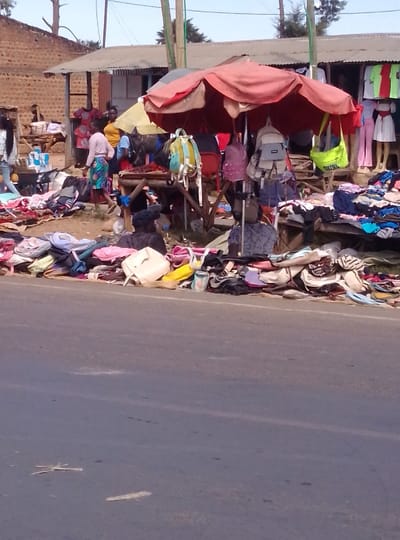 The scene depicts a bustling roadside market in South Kabras ward, Kenya, viewed from across an asphalt road. In the foreground, the paved road surface shows faint white lines and minor debris. The midground features a market stretching along the roadside, characterized by numerous makeshift stalls. A prominent, large reddish-orange market umbrella shades a central display area where various backpacks and handbags are suspended from wooden frames.
Piles of clothing, shoes, and other merchandise are laid out directly on mats or low wooden tables adjacent to the road. At least six individuals are visible within the market. One person in a light top and patterned skirt walks leftward near the center. Others are standing or seated amidst the goods, appearing to be vendors or shoppers. Behind the primary market area, further stalls with corrugated metal roofs display additional clothing items, some on mannequins, in various bright colors. In the background, brick buildings with similar metal roofs are visible, backed by trees and utility poles under a clear sky. The overall lighting suggests daylight conditions.