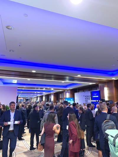 This image depicts a crowded indoor professional event, likely a conference or forum, held at a large convention facility, identified as **EXPO XXI Warszawa** in Warszawa, Poland.

The scene features a wide hall or corridor with a high white ceiling, accented by numerous circular recessed spotlights and prominent linear blue LED strip lighting running along recessed architectural details. Walls are panelled, displaying light-colored sections interspersed with darker, possibly wood-finished, horizontal bands. The floor is covered with a patterned carpet in light beige or off-white tones, adorned with intricate, curvilinear dark brown or beige designs.

Approximately 50 to 100 individuals, predominantly adults, are visible throughout the space, standing, conversing, and moving. Most attendees are dressed in business or business-casual attire, including dark suits, blazers, white shirts, and business dresses in various colors such as dark red/burgundy. Many wear lanyards with ID badges.

In the left foreground, a middle-aged man with short grey hair, wearing a dark blue suit and a white shirt, holds two white cups, possibly containing coffee or tea. He looks towards the right, appearing to navigate the crowd.

In the mid-ground and background, the crowd is densely packed, suggesting active networking. Several people are seen holding beverages or small personal bags. A woman in the left-center wears a dark red dress and holds a brown handbag. Another woman further right wears a dark red suit. A man in the far right background has a light green/grey backpack.

In the background, two large digital display screens and a white banner are visible. The central white banner displays text including "MOBILIS WORLD FOR UKRAINE" and "FC-UP." To its right, a blue-lit screen prominently displays "WELCOME" at the top, followed by "ECONOMIC FORUM" and other graphic elements below. The overall atmosphere is bustling and professional.