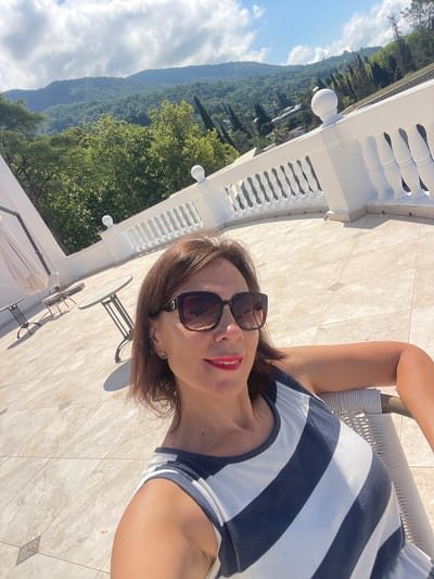 A woman is seated on a chair on a patio with a view of mountains. The woman has brown hair and is wearing sunglasses and a striped shirt. The patio has a tiled floor and a white railing. In the background, there are lush green mountains and trees. The sky is partly cloudy.