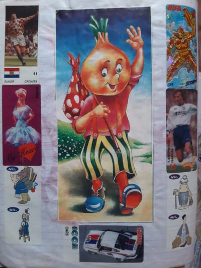 The image displays a collection of stickers and a large, colorful illustration of a cartoon onion character on a piece of paper. The paper appears to be a page from a scrapbook or album, with several other smaller stickers arranged around the central illustration.
On the left side of the page, there are three rectangular stickers. The top one shows a soccer player in a Croatian jersey celebrating, with the Croatian flag and the name "SUKER" and "CROAITA" below. Below this is a sticker of a doll in a blue dress with "My Heart" written on it. The bottom left sticker is a cartoon of a man with a backpack.
To the right of the central onion illustration are several more stickers. The top one features a metallic, armored figure with a weapon, labeled "BIFA" and "STORM". Below this is a sticker of a soccer player in a white and blue jersey, identified as "T.SHERWOOD" and "ISKOÇIA". Further down, there are two more small cartoon stickers: one of an elderly woman with a cane and shopping bag, and another of a man in a suit carrying luggage.
The central illustration is of a cheerful, anthropomorphic onion wearing a red shirt, suspenders, and striped green and yellow pants. It carries a red and white polka-dotted bag over its shoulder and appears to be waving with one hand. The background of the illustration is a blue sky with green hills.
At the bottom of the page, near the center, is a sticker depicting a race car with the number "65" and the word "DIXIE" visible. There are also vertical text labels like "CARS" and "BOOM" on this sticker.
The overall scene is that of a personal collection of varied stickers, likely gathered by an individual. There is no identifiable information within the image that would suggest a specific location in the city of Semey, Kazakhstan.