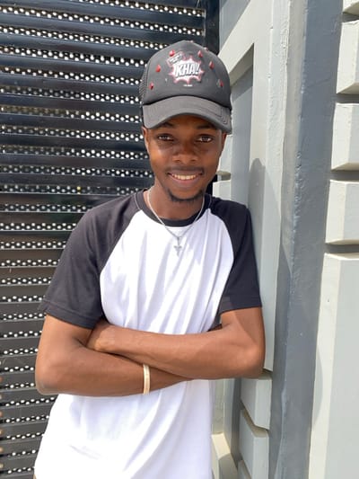 A young male subject is depicted outdoors in Joma, Ghana, facing the camera with a smile. He wears a black baseball cap featuring a "KHA!" speech bubble graphic and red drop-like embellishments, a black and white raglan-style t-shirt, a silver-colored chain with a cross pendant, and a light-colored bracelet on his right wrist. His arms are crossed over his chest. The background on the left consists of a dark, horizontally slatted structure. On the right, the subject leans against a light grey concrete wall with horizontal protrusions. The scene is illuminated by direct, bright natural light.