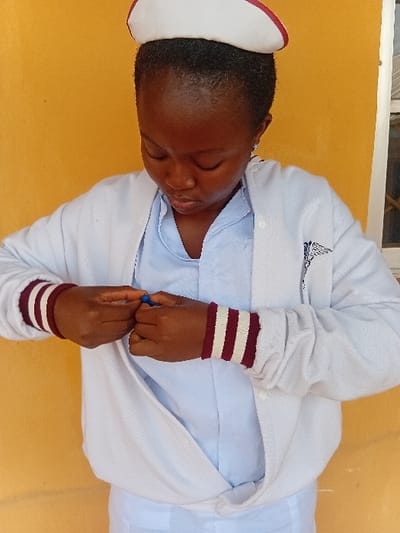 A young Black female is depicted from the upper torso, standing against a plain mustard yellow wall in Ejule, Nigeria. She wears a white nurse's cap with a red band, a light blue uniform shirt, and an open white long-sleeved jacket. The jacket features maroon and white striped cuffs, with a dark emblem, possibly a medical symbol, visible on the left chest. Her head is tilted downwards, eyes focused on her hands, which are clasped at her waist, manipulating a small blue object. A partial view of a white-framed window or structure with dark horizontal bars is visible on the far right of the background.