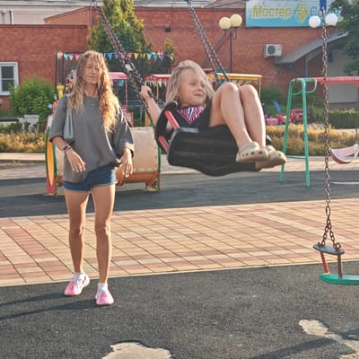 In the city of Kurganinsk, Russia, an outdoor playground scene captures a mother and her young daughter. The mother, a woman with long, curly blonde hair, stands on the paved area, wearing a grey t-shirt, denim shorts, and pink shoes. She is holding a light blue bag over her shoulder and has a watch on her left wrist. Her daughter is actively engaged on a swing, her legs and feet visible as she enjoys the motion. She is wearing a black shirt with pink zebra-striped trim and beige crocs. The playground features a swing set with metal chains, a black swing seat, and a green and red swing seat. In the background, a brick building with signage that reads "Мастер" is visible, along with trees and other playground equipment including a red toy truck and a pink toy car. The ground is a combination of dark grey rubberized surfacing and a patterned paved area.