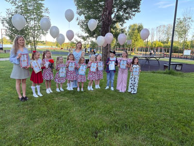 An outdoor scene in a public park in Zaprudnya, Russia, depicts a group of two adult women and ten children posing together. The foreground is covered in green grass. In the middle ground, from left to right, a young blonde woman wearing a light blue collared shirt and a patterned white and green skirt holds a framed certificate and a white balloon string. To her right, a line of ten children, predominantly girls, stands facing the camera. The first four girls from the left wear matching red and white strawberry or cherry-patterned dresses. The third girl from the left holds a small bouquet of red roses in addition to a framed certificate. Another young blonde woman, wearing a light blue dress, stands slightly behind the fourth and fifth girls, also holding a white balloon string.

All children and the first adult woman are holding framed certificates, mostly displaying the word "Грамота" (Gramota) in Russian. The children's attire varies towards the right: a boy in a plaid shirt, jeans, and a baseball cap stands next to a girl in a light pink top and trousers, who stands next to a girl in black and white patterned trousers and a black top.

Above the group, several white and light pink balloons float, tethered by strings. In the background, numerous green-leafed trees are visible. To the left, a basketball hoop structure is partially seen. Further in the background, a body of water is visible, with more trees and structures beyond it. A public recreation area is evident, featuring parallel bars, other exercise equipment, a table tennis table, and benches. Several individuals are present in the distant background, interacting with the recreational facilities. The sky is clear blue with minimal cloud cover.