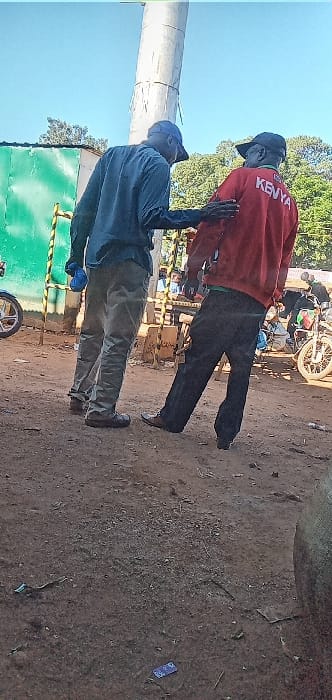 The image shows two men standing in a dusty, open-air environment.  The man on the left is wearing a blue shirt, khaki pants, and a blue hat. The man on the right is wearing a red sweatshirt with the word "KENYA" written on the back, black pants, and a black hat. They appear to be having a conversation and are gesturing with their hands. There is a tall, white pole in the background. The environment appears to be a casual outdoor setting. The coordinates, 0.562 latitude and 34.805 longitude, place the location in the town of Kivaywa, Kenya.