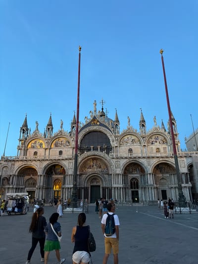 This image depicts St. Mark's Basilica and a portion of St. Mark's Square in Venice, Italy, at dusk or early evening. The prominent Byzantine facade of the basilica dominates the background, featuring multiple arches adorned with intricate carvings, statues, and glowing golden mosaics depicting religious scenes. Several spires and architectural details rise above the main structure, contributing to its ornate appearance.
Framing the basilica in the mid-ground are two tall, slender flagstaffs, painted a deep red, each topped with a golden, winged lion finial, the symbol of Venice. The square itself is paved with dark gray flagstones and is populated by numerous people. In the foreground, with their backs to the camera, are several individuals: a person wearing a white t-shirt, yellow shorts, and a blue backpack, looking towards the basilica; to their left, three women are walking, one with long dark hair, a dark top, and black leggings, another in a denim jacket, green pleated skirt, and white cross-body bag, and the third in a dark top and white shorts.
Further into the square and closer to the basilica, many other pedestrians are visible, moving about or standing. On the far left of the frame, a mobile vendor's cart, laden with apparel, is situated near the basilica's lower arches. The sky above is a clear, deep blue, contrasting with the illuminated architectural elements of the basilica.