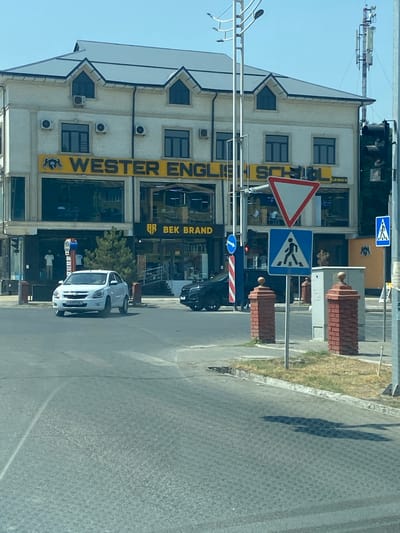 A white sedan is stopped at an intersection in Andijan City, Uzbekistan. The car is facing right, with its front wheels turned slightly to the right. Behind the car is a building with a sign that reads "WESTERN ENGLISH SCHOOL". The building has several windows and air conditioning units. There are traffic lights and pedestrian crossing signs at the intersection. The sky is blue and clear.