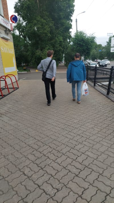 Two people are walking on a pedestrian sidewalk paved with interlocking concrete blocks. The person in the foreground is wearing a grey textured jacket and black pants, with a dark bag slung across their shoulder. The person behind them is wearing a blue jacket and blue jeans, carrying a white plastic shopping bag. Both individuals appear to be walking away from the camera.
The scene is outdoors, with lush green trees lining the left side of the sidewalk. A black metal fence runs along the right side of the sidewalk, separating it from what appears to be a road or parking area where several cars are visible. Overhead, the sky is overcast, suggesting a cloudy day.
On the left side of the image, partially visible, is a building with a sign. The sign has a blue and red circular logo with a white stripe and some red lines. Below this, there's a yellow sign with a white arrow pointing to the right, and the word "SAVAGE" written in white text. There is also text in what appears to be Cyrillic on the left side of the yellow sign. The overall impression is a typical urban or suburban street scene on a cool, cloudy day. No specific emotions are discernible from the subjects.