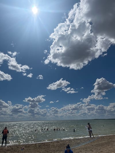 This is a vertical, eye-level, outdoor, full shot photograph taken on a sunny day at a beach. The foreground consists of a sandy shore with a few scattered objects, including a pair of pink flip-flops. The middle ground is a body of water, likely a lake, with numerous individuals swimming and wading. The water is shimmering from the sunlight, and there are gentle waves lapping at the shore. The background features a vast expanse of blue sky dotted with fluffy white and grey clouds. The sun is prominently visible in the upper left quadrant of the image, casting a bright glare. Two individuals are visible standing on the shore in the foreground, one in the lower left wearing an orange life vest and a hat, facing the water, and another in the lower right, a woman wearing a swimsuit and holding a floatation device. The location is identified as Gres-2, Kazakhstan.