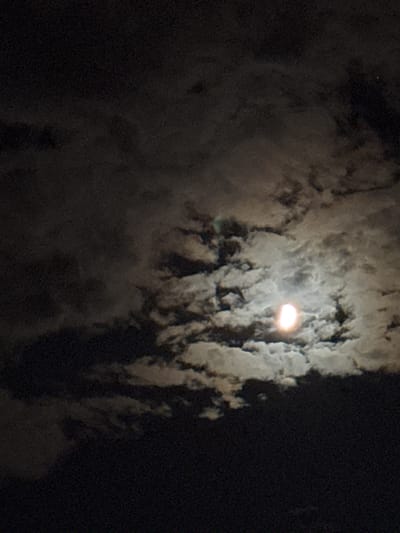 The image depicts a night sky over Rafailovići, Montenegro. A bright, semi-illuminated moon is visible, partially obscured by wispy clouds. The clouds vary in density, with some appearing dark and heavy, while others are lighter and translucent, allowing the moonlight to shine through. The overall impression is one of a slightly overcast night, with the moon's glow creating a soft, ethereal light. The lower portion of the image transitions to a dark, possibly terrestrial area, which is not clearly defined. There are no people or specific landmarks visible that would indicate a precise location within Rafailovići.